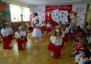 Na tle dekoracji z mapą Polski i dziećmi z chorągiewkami przedszkolaki tańczą w parach. Jedna dziewczynka na środku śpiewa.